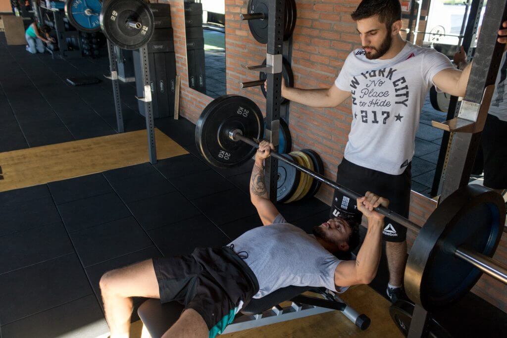 Man lifting weights on a bench press with assistance from a spotter, highlighting potential risks of guidance from the wrong personal trainer.